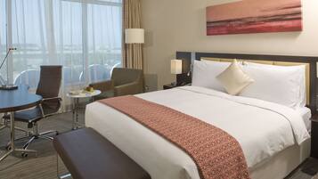 Habitación de hotel con una cama grande, un escritorio, una silla, una mesita redonda y vistas de la ciudad a través de grandes ventanales.