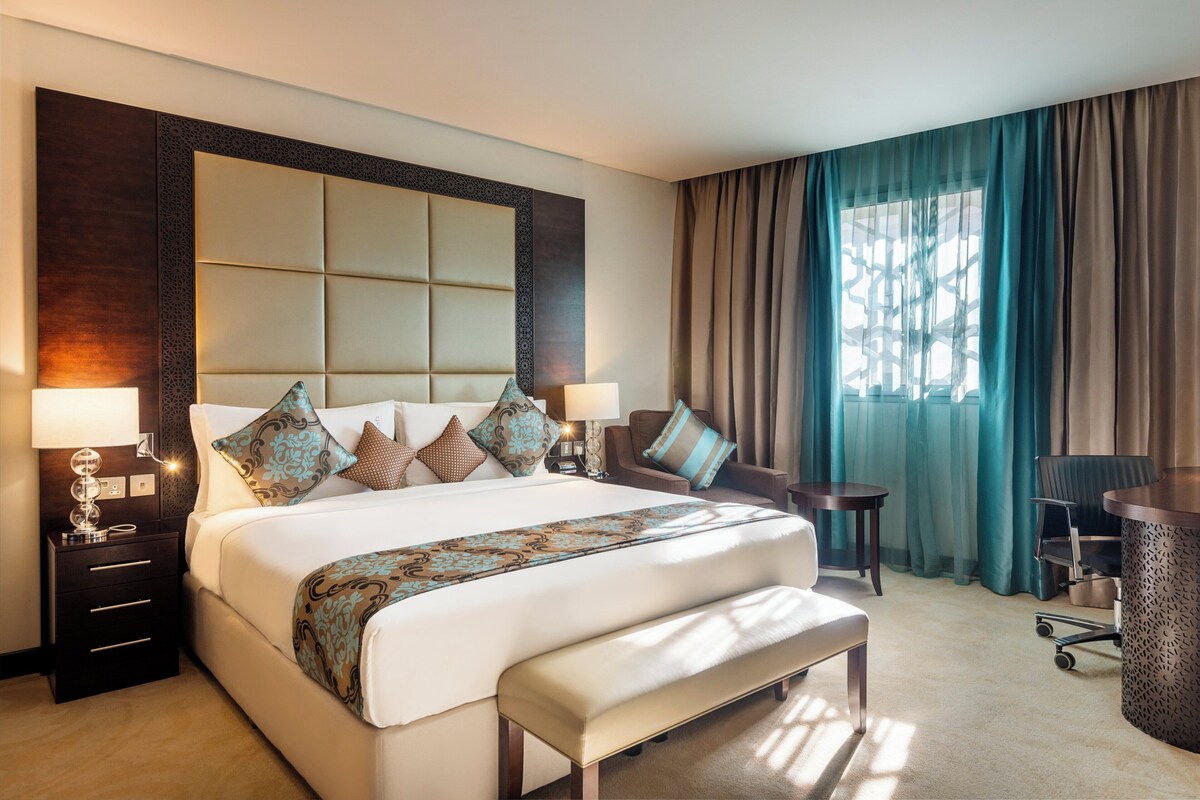 A hotel room with a large bed, a desk, and a chair. There is a window with curtains and a lamp on the side table.