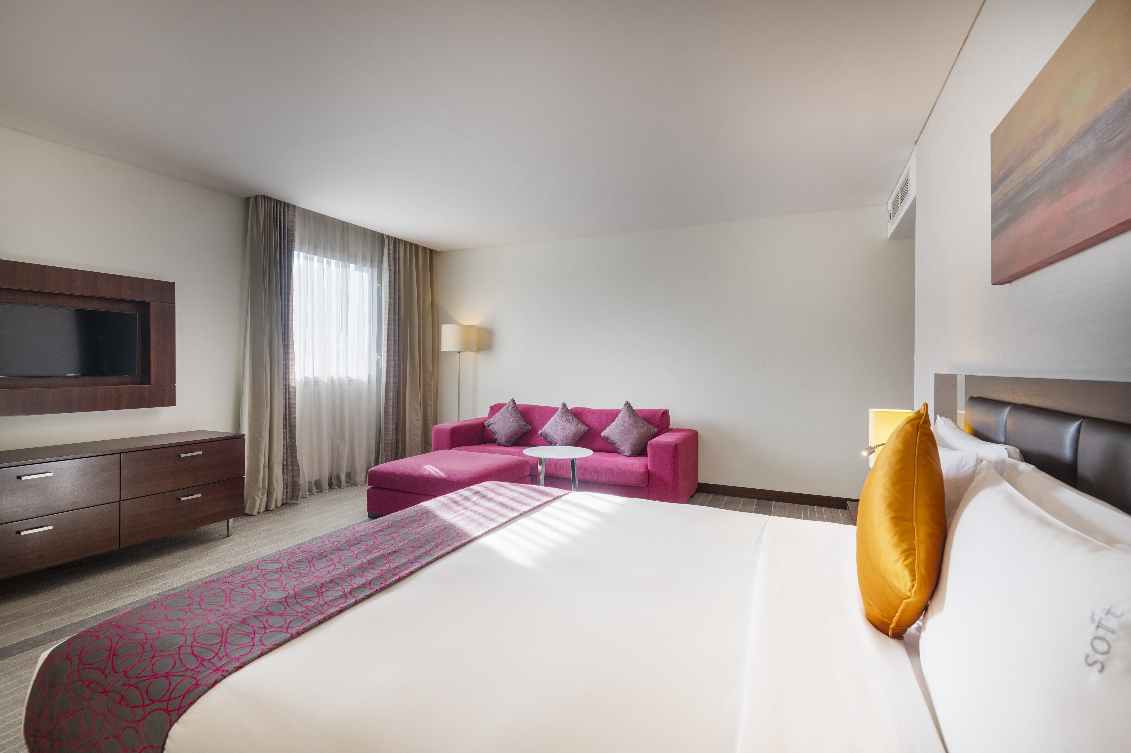 A hotel room with a large bed, a pink sofa, a flat-screen TV, and a window with curtains.