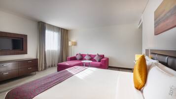 A hotel room with a large bed, a pink sofa, a flat-screen TV, and a window with curtains.