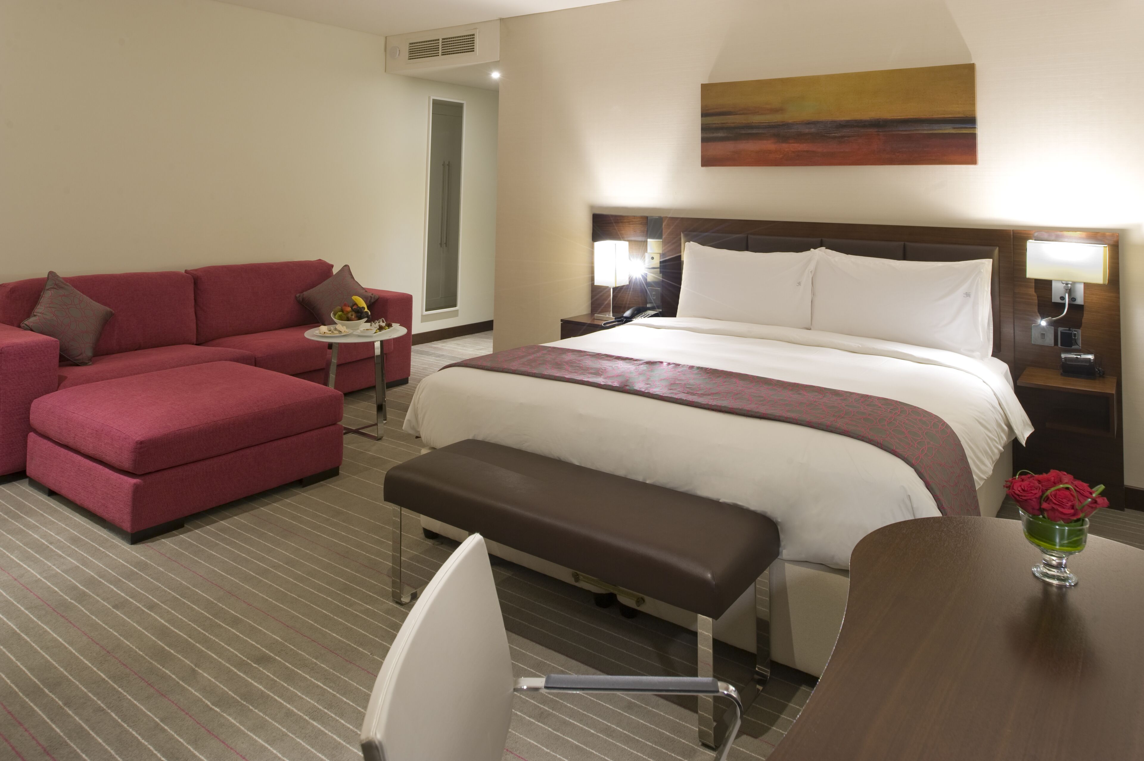 Quarto de hotel com uma cama grande, um sofá vermelho, uma pequena mesa com abajur e um banquinho.