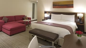 Quarto de hotel com uma cama grande, um sofá vermelho, uma pequena mesa com abajur e um banquinho.