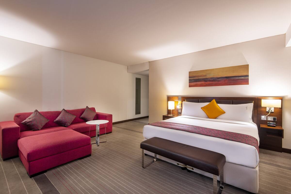 A hotel room with a large bed, a red sofa, a small table, and a bench.