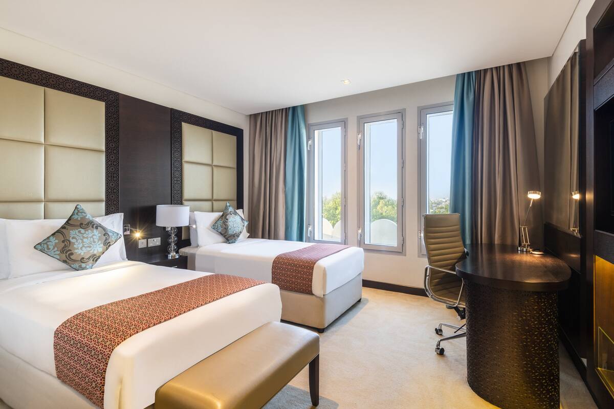 A hotel room with two beds, a desk, a chair, and large windows.