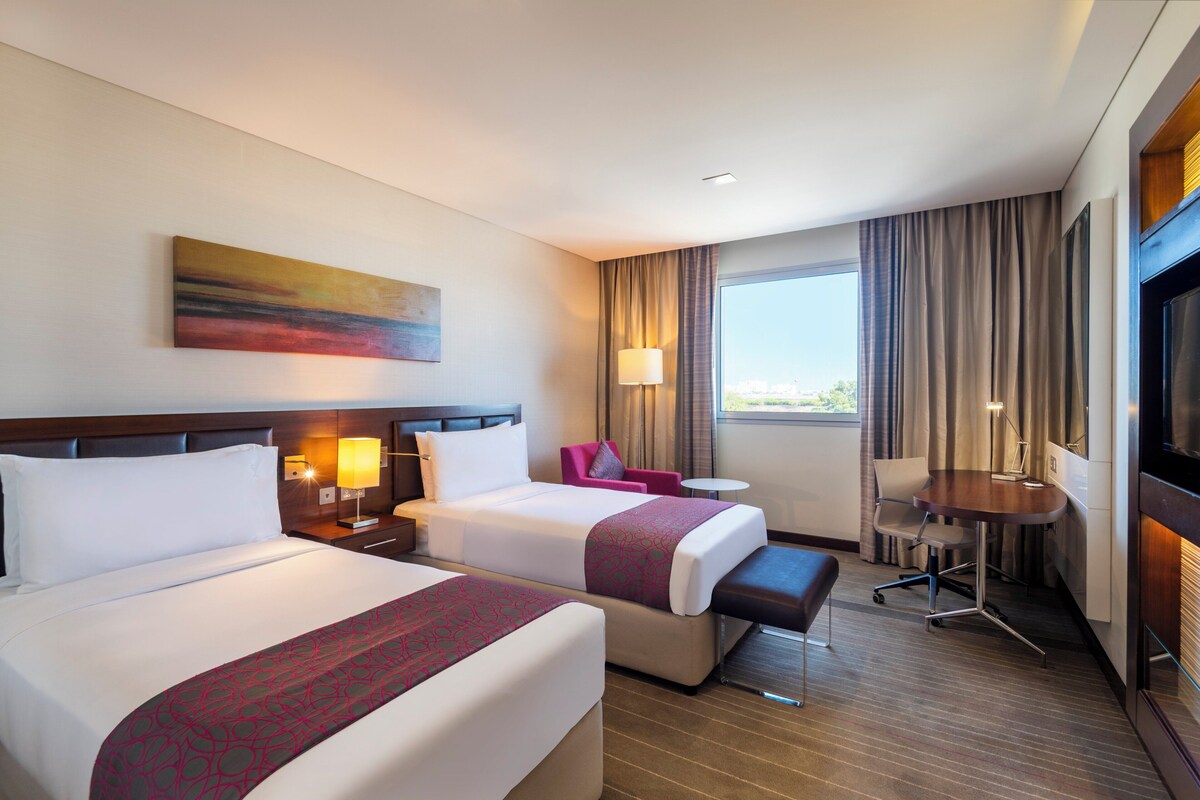 A hotel room with two beds, a desk, a chair, a small table, and a TV.