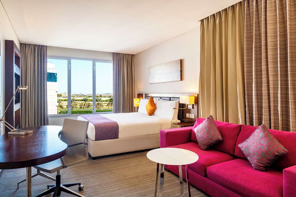 A hotel room with a bed, a red sofa, a small white table, a desk, and a large window with curtains.