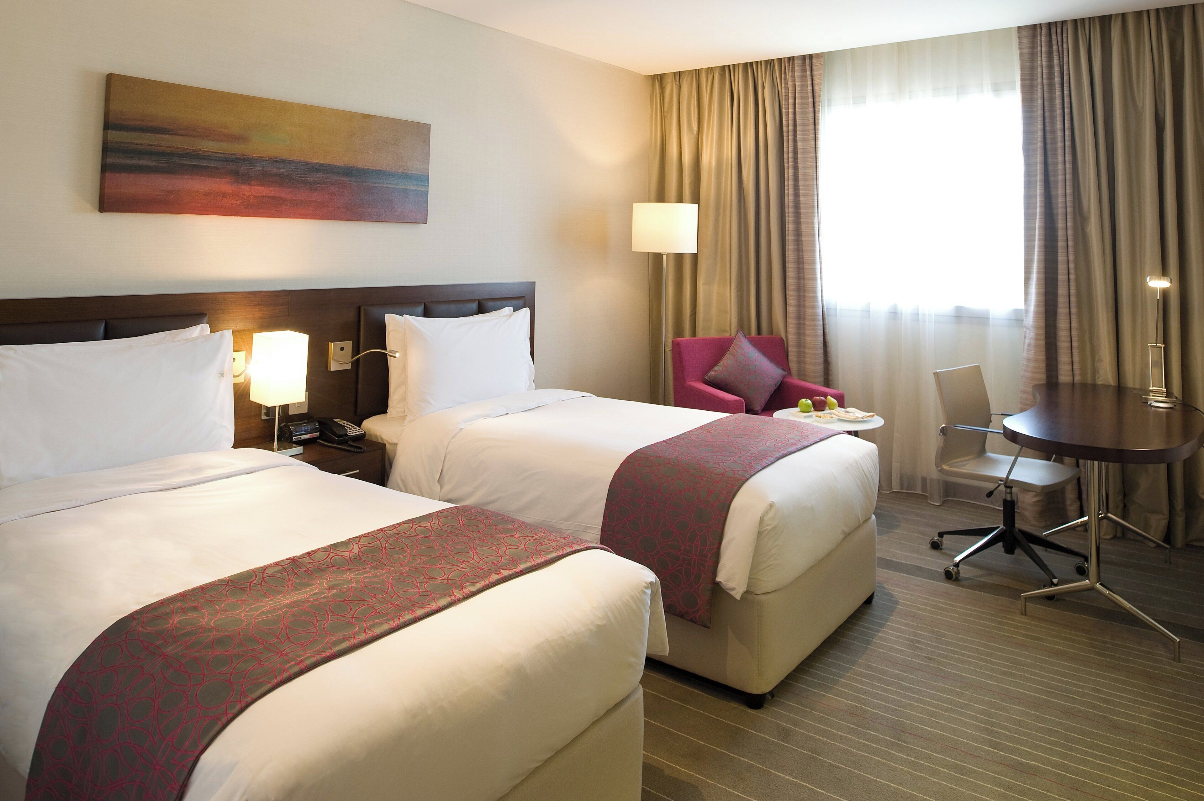 A hotel room with two beds, a desk, a chair, a lamp, and a painting on the wall.