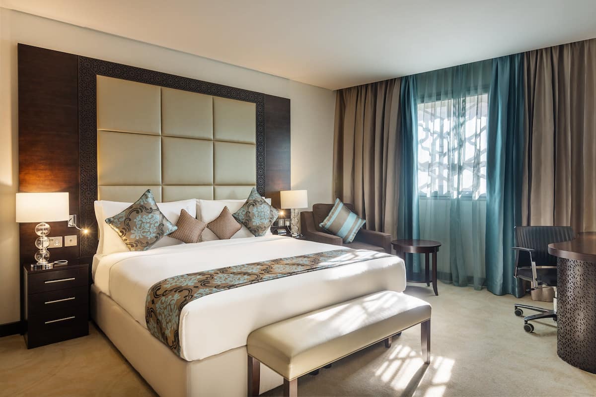 A hotel room with a large bed, a desk, and a chair. There is a window with curtains and a lamp on the side table.