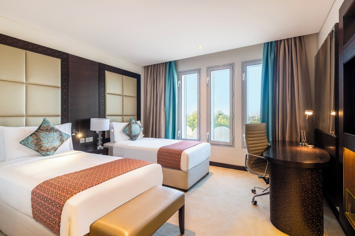 A hotel room with two beds, a desk, a chair, and large windows.