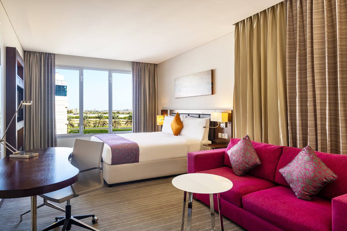 A hotel room with a bed, a red sofa, a small white table, a desk, and a large window with curtains.