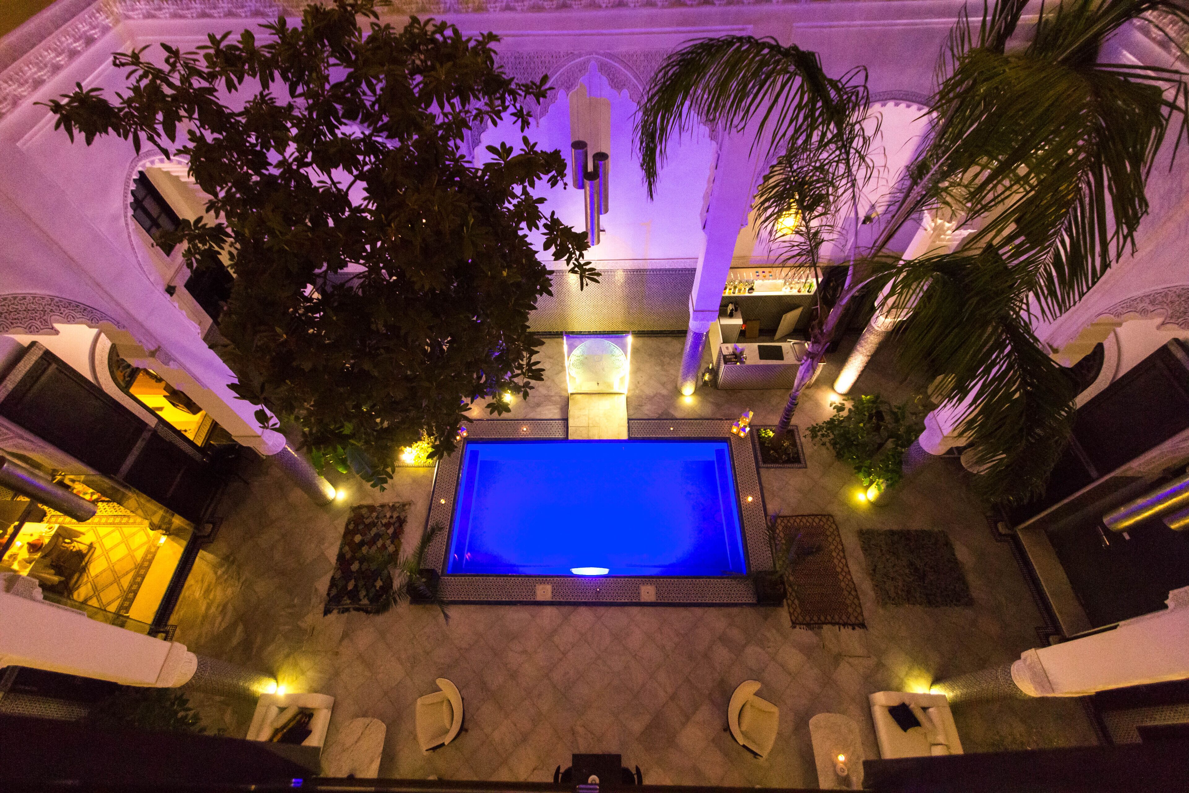 A rooftop pool area with a blue pool, palm trees, and a seating area with chairs and a table.