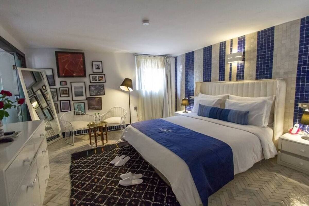 A hotel room with a large bed, a bedside table, a chair, a desk, and a mirror. There are framed pictures on the wall and a window with curtains.