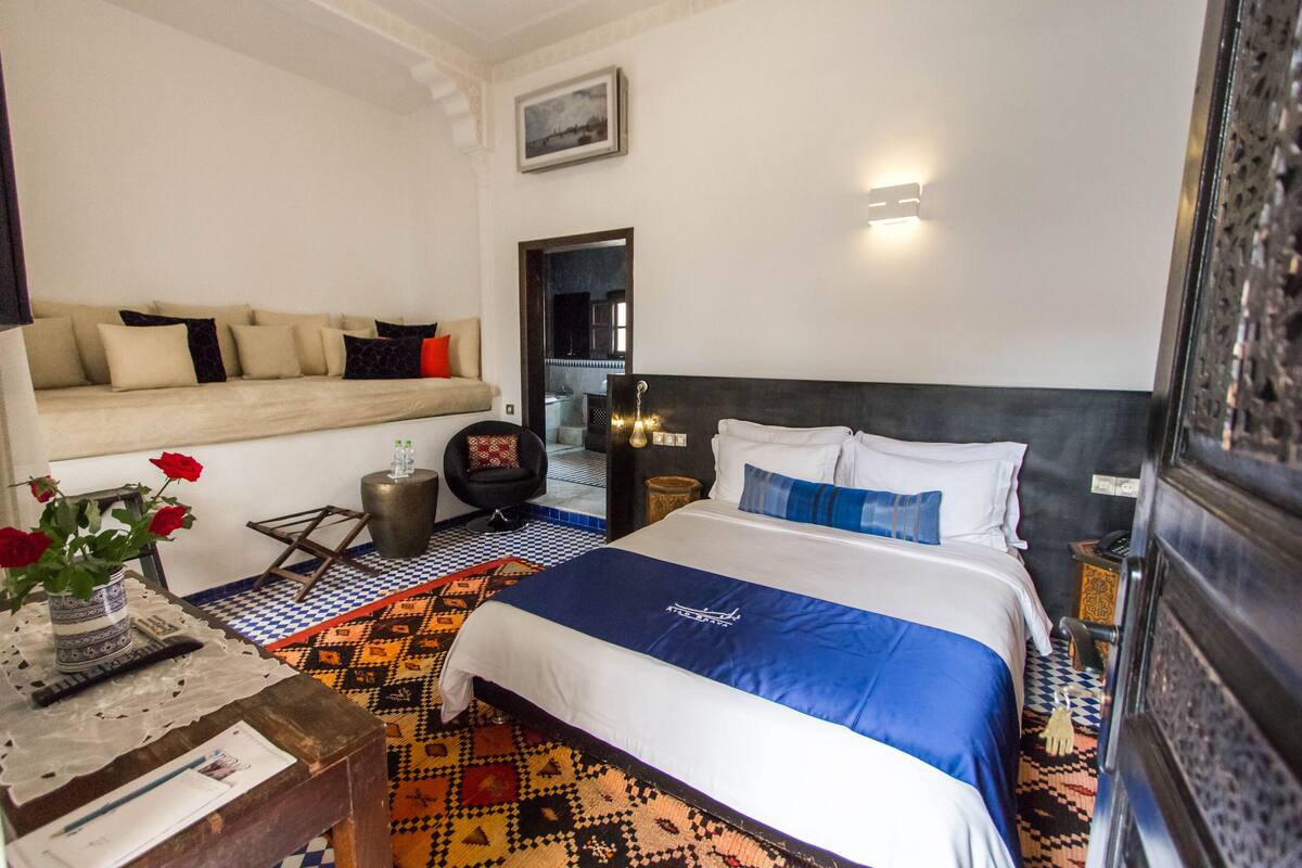 A hotel room with a bed, a sofa, a small table with a vase of flowers, and a patterned rug.
