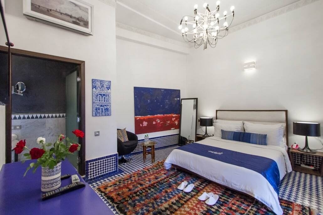 Una habitación de hotel con cama, una silla, una mesa con un jarrón de flores, una alfombra y un candelabro.