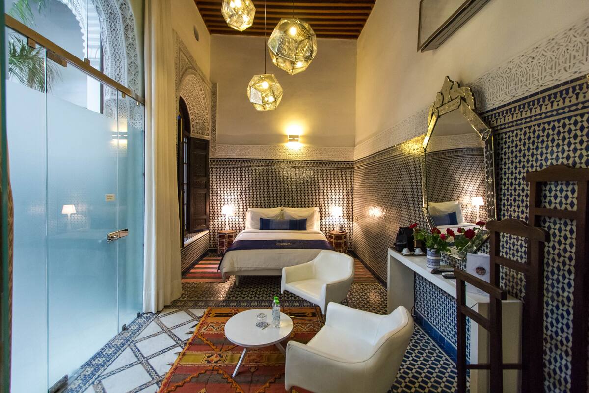 A hotel room with a bed, two chairs, a small table, a mirror, and patterned walls.