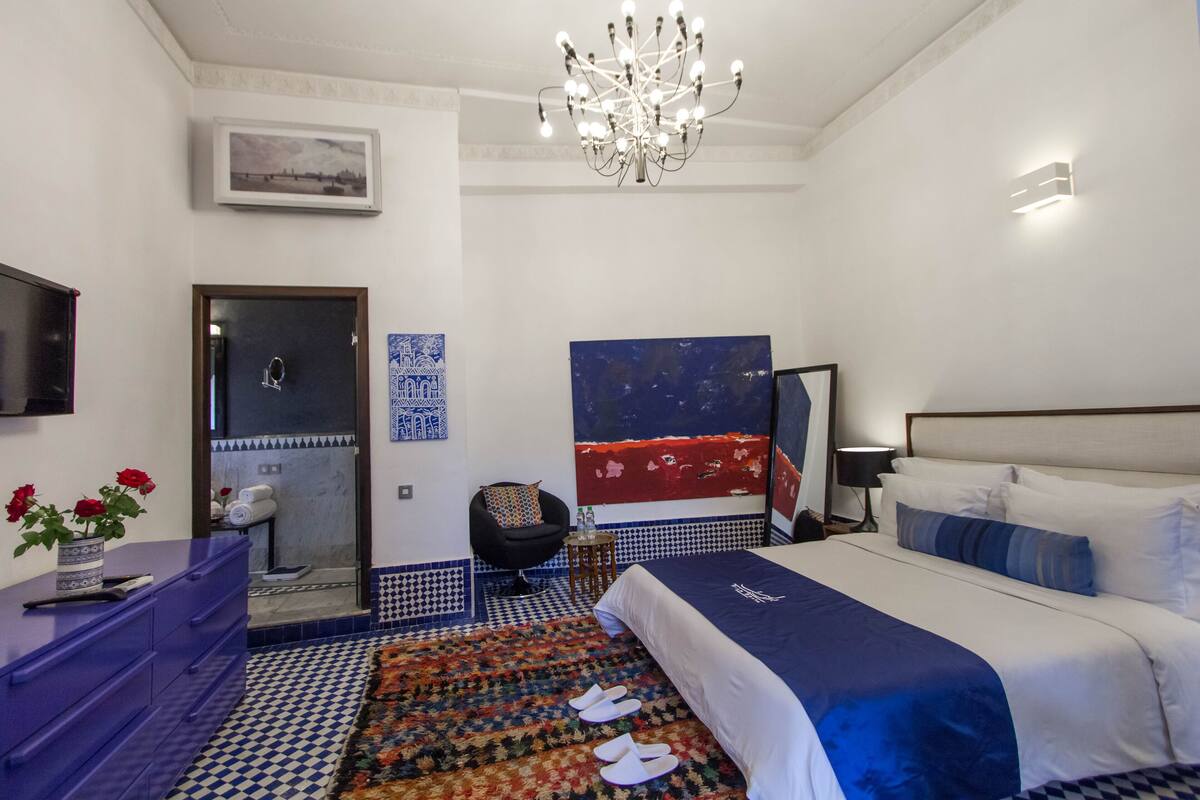 A modern hotel room with a large bed, a chandelier, a bathroom with a shower, and a blue and white checkered floor.