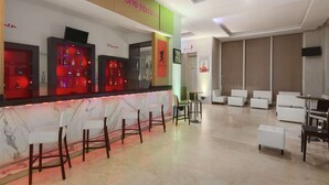 Un área de bar moderna con encimera de mármol, varios taburetes y un televisor. Se observan sillas y mesas blancas al fondo.