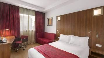 Une chambre d’hôtel avec un grand lit, un bureau avec une chaise, un canapé rouge et une tête de lit en bois.