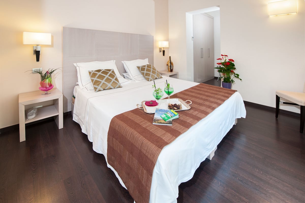 A neatly made bed with a brown blanket, two bedside tables with lamps, a small potted plant, and a magazine on the bed.