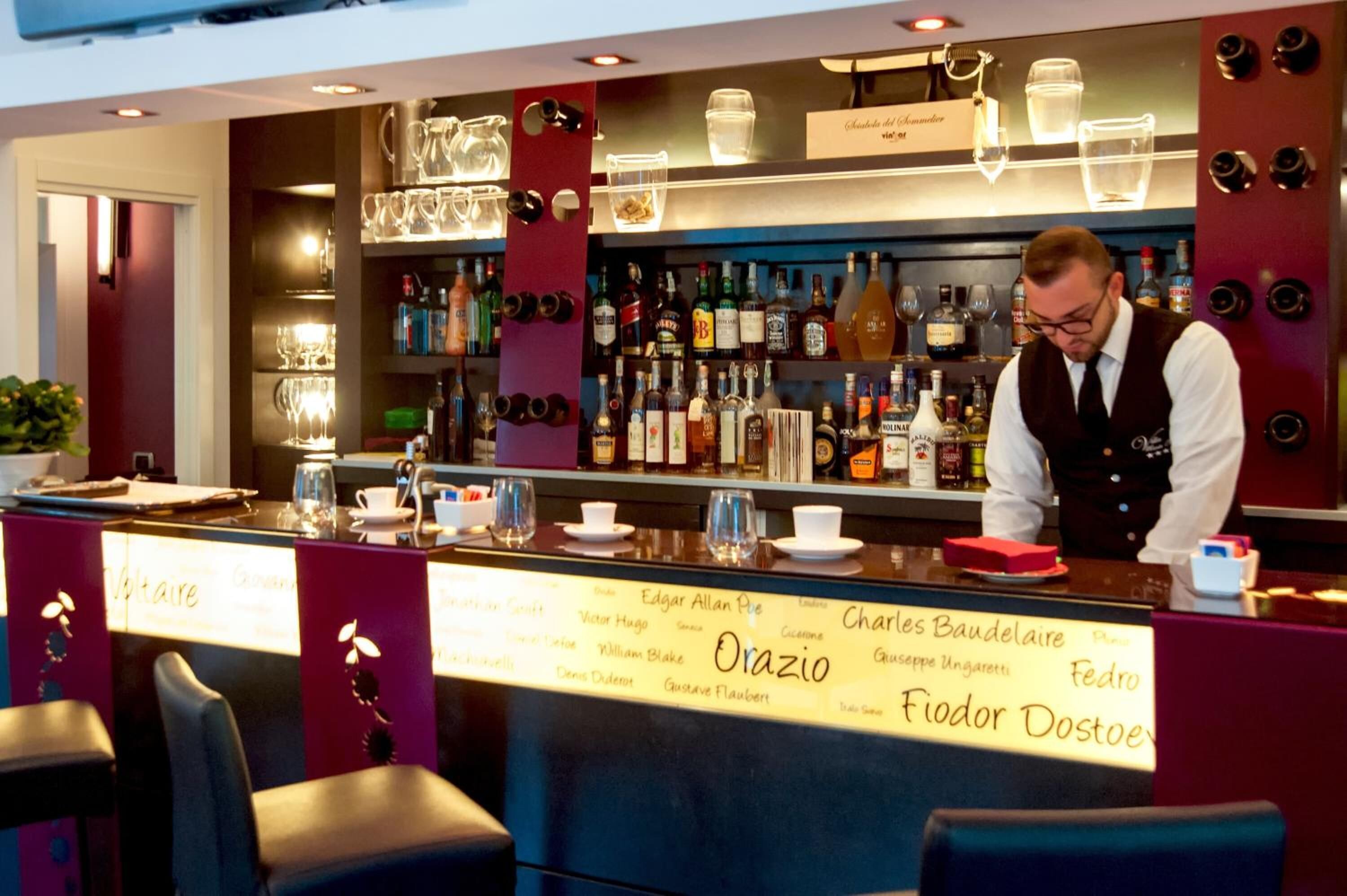 A bar area with a bartender, shelves stocked with bottles, glasses, and a menu board listing names.