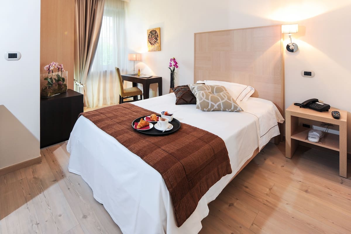 A hotel room with a bed, bedside table, lamp, and a tray with croissants and coffee.
