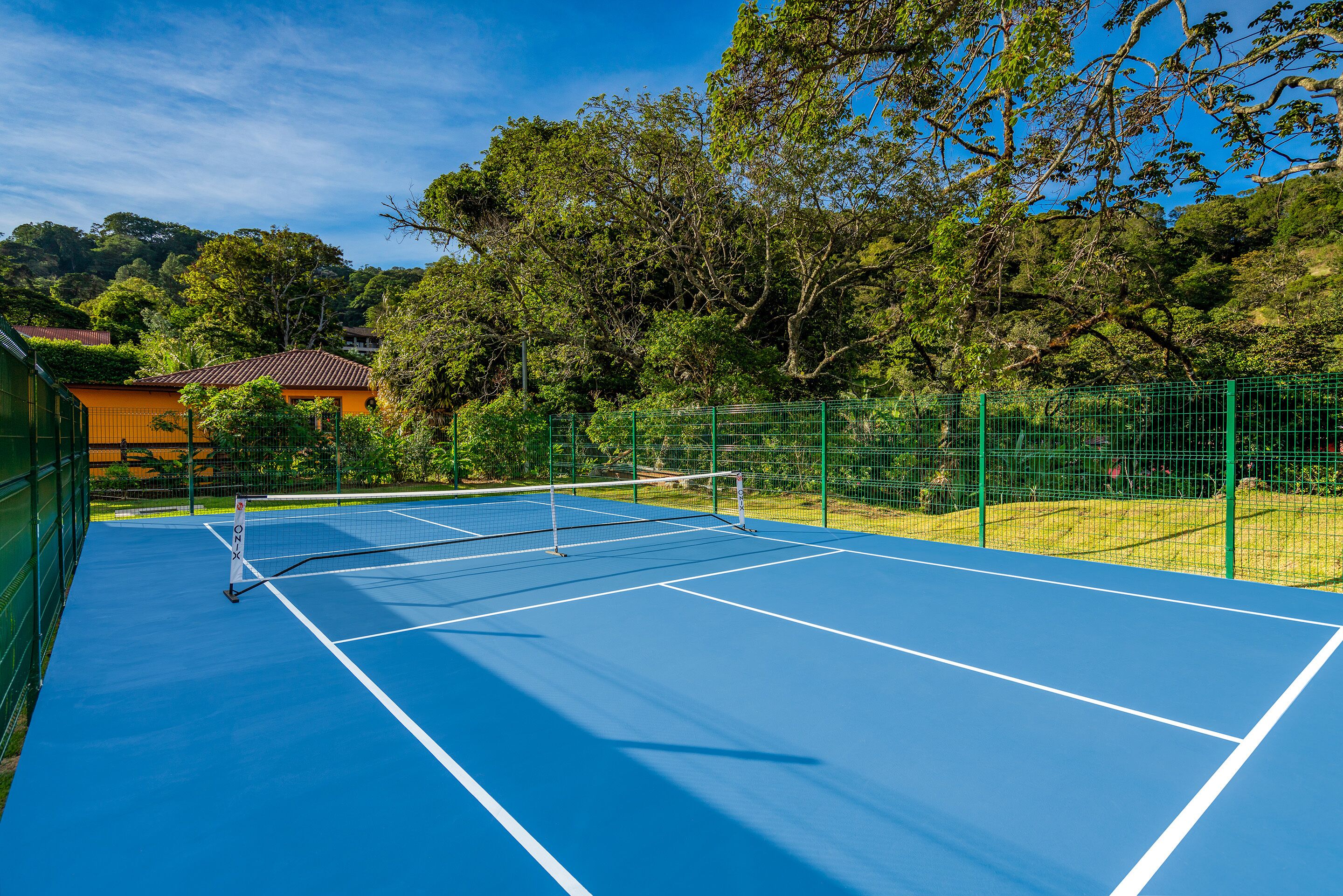 Una cancha de tenis al aire libre con red, rodeada de vallas verdes y árboles.