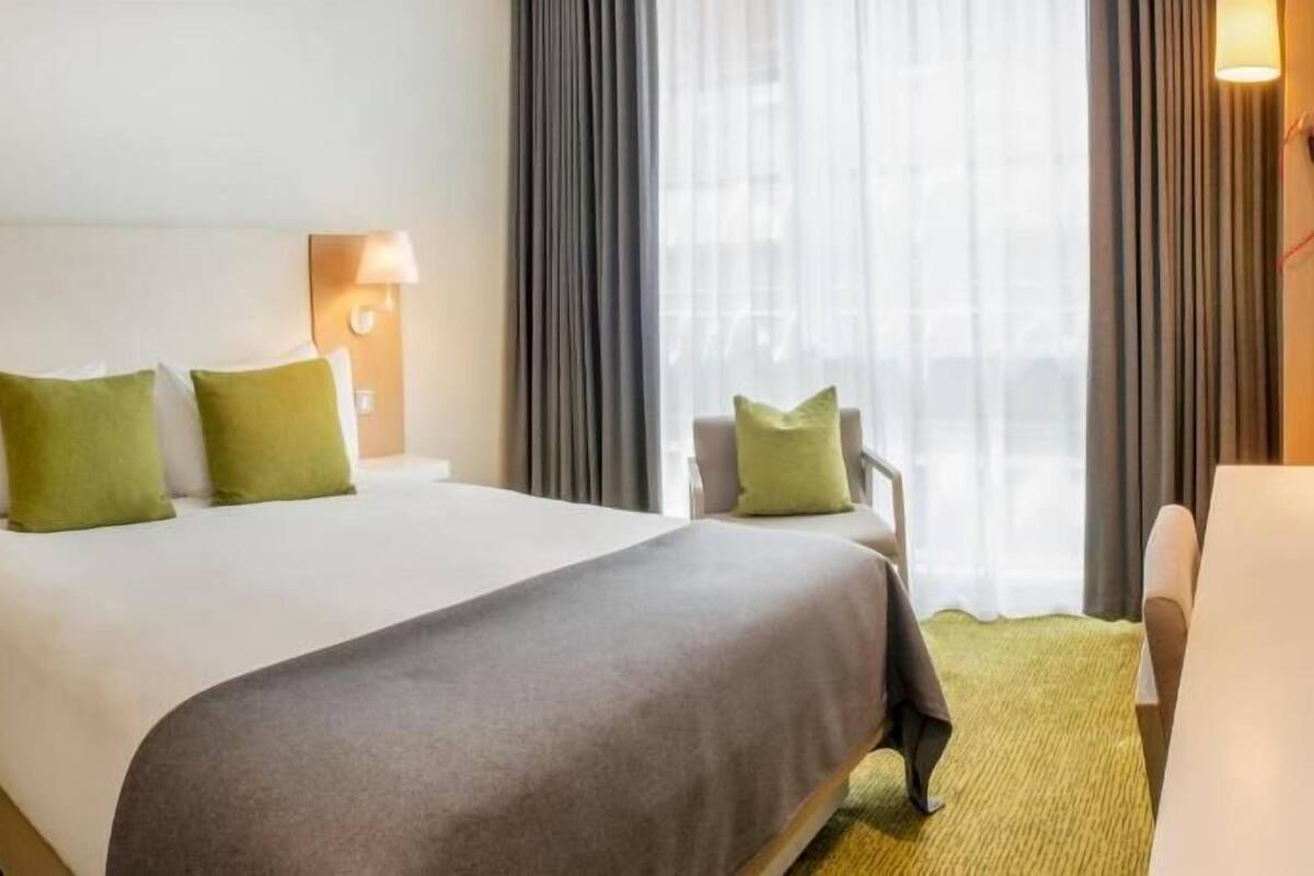 Hotellihuone, jossa on suuri sänky, kaksi yöpöytälamppua, tuoli ja ikkuna, jossa on verhot.