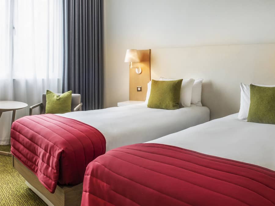 A hotel room with two beds, each with a red bedspread and green pillows, a small table, and a chair. There are wall-mounted lamps on either side of the beds.