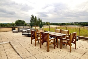 A patio with a wooden table and chairs, surrounded by a low fence and greenery.