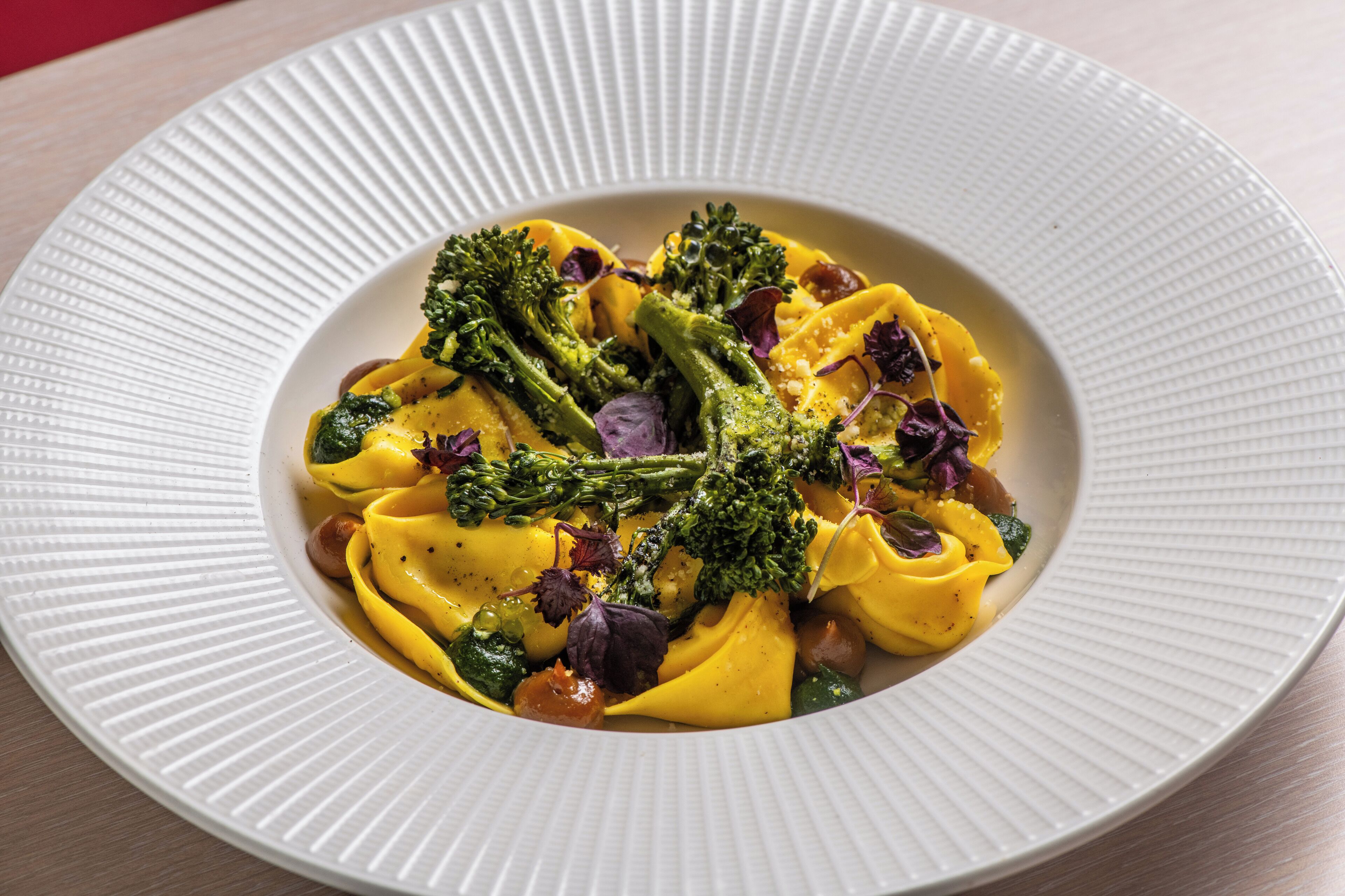 A white plate with a textured edge, containing a pasta dish with broccoli and purple leaves, possibly microgreens, on a wooden table.