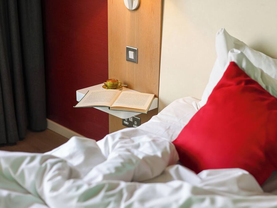 A neatly made bed with white bedding and a red pillow, a bedside table with an open book, and a wall-mounted light.