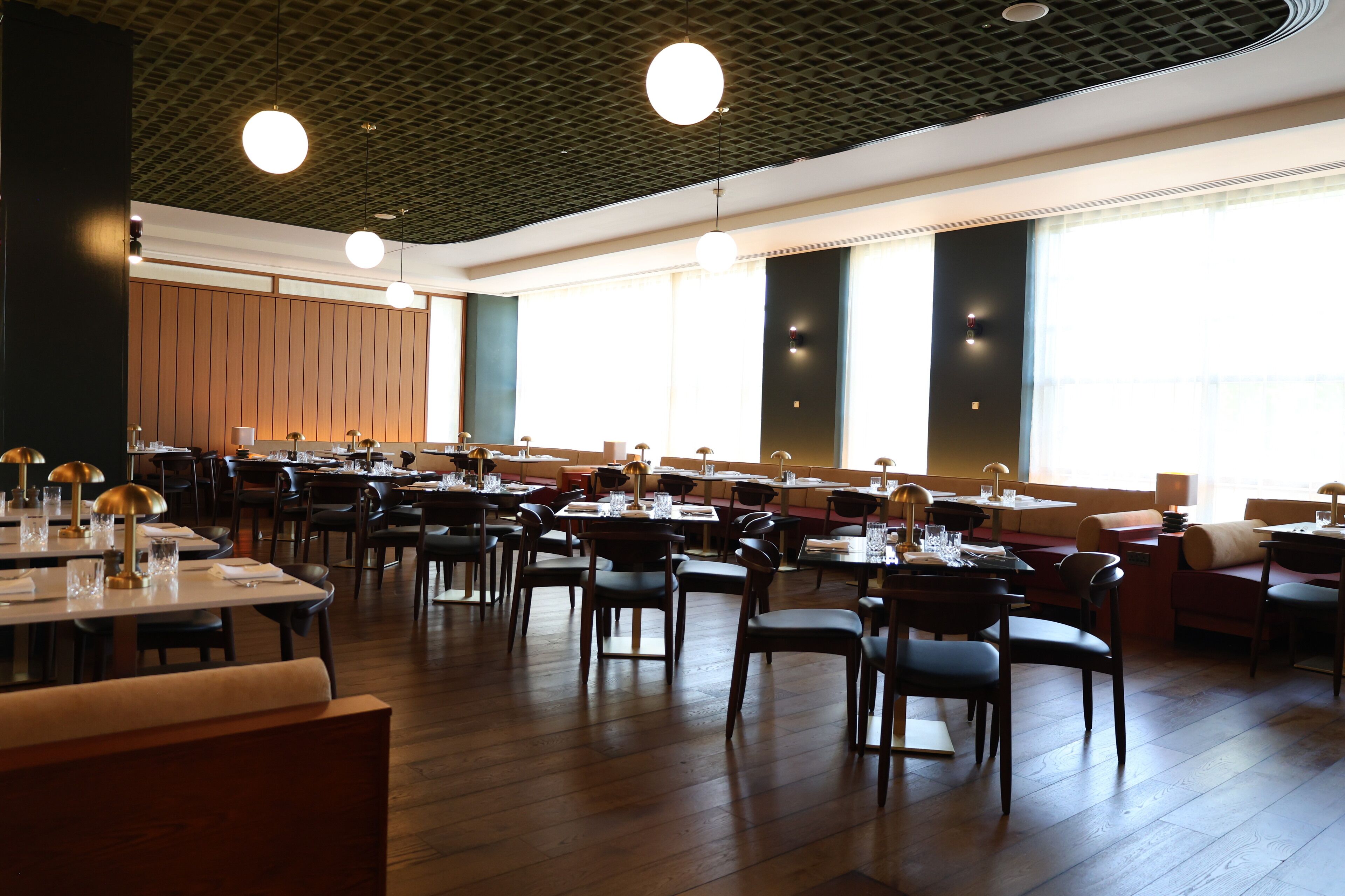 A spacious restaurant with multiple dining tables and chairs, large windows, and a modern ceiling design.