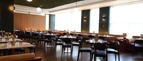 A spacious restaurant with multiple dining tables and chairs, large windows, and a modern ceiling design.