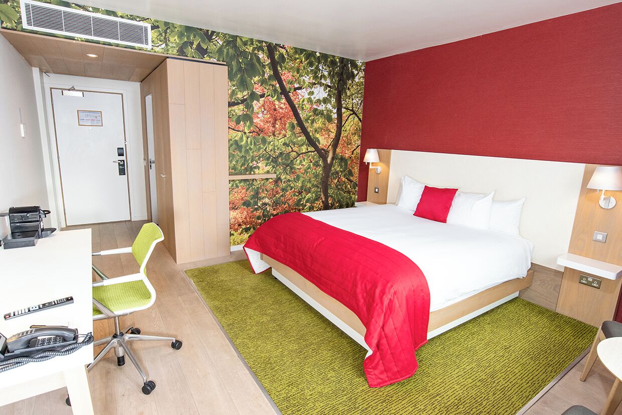 A hotel room with a bed, desk, chair, and a wall with a tree mural.