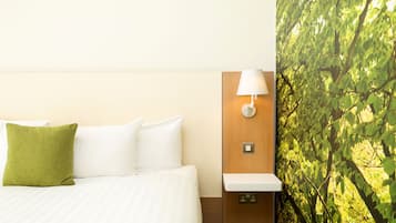 Habitación de hotel con una cama, almohadas blancas, una almohada verde, una lámpara de pared y una pared con vistas a un árbol frondoso.