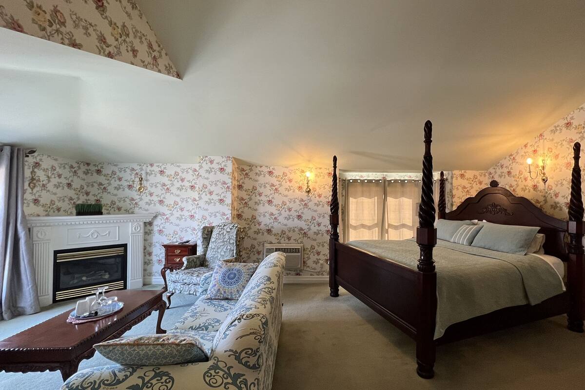 A four-poster bed with a wooden headboard, a sofa with patterned upholstery, a small table with a vase, and a fireplace with a mantel.