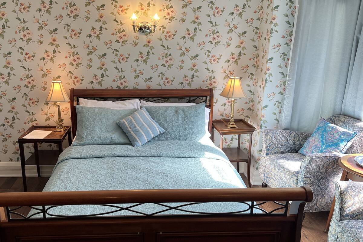 A neatly made bed with a floral-patterned bedspread and pillows, flanked by two bedside tables with lamps, in a room with floral wallpaper and a patterned armchair.