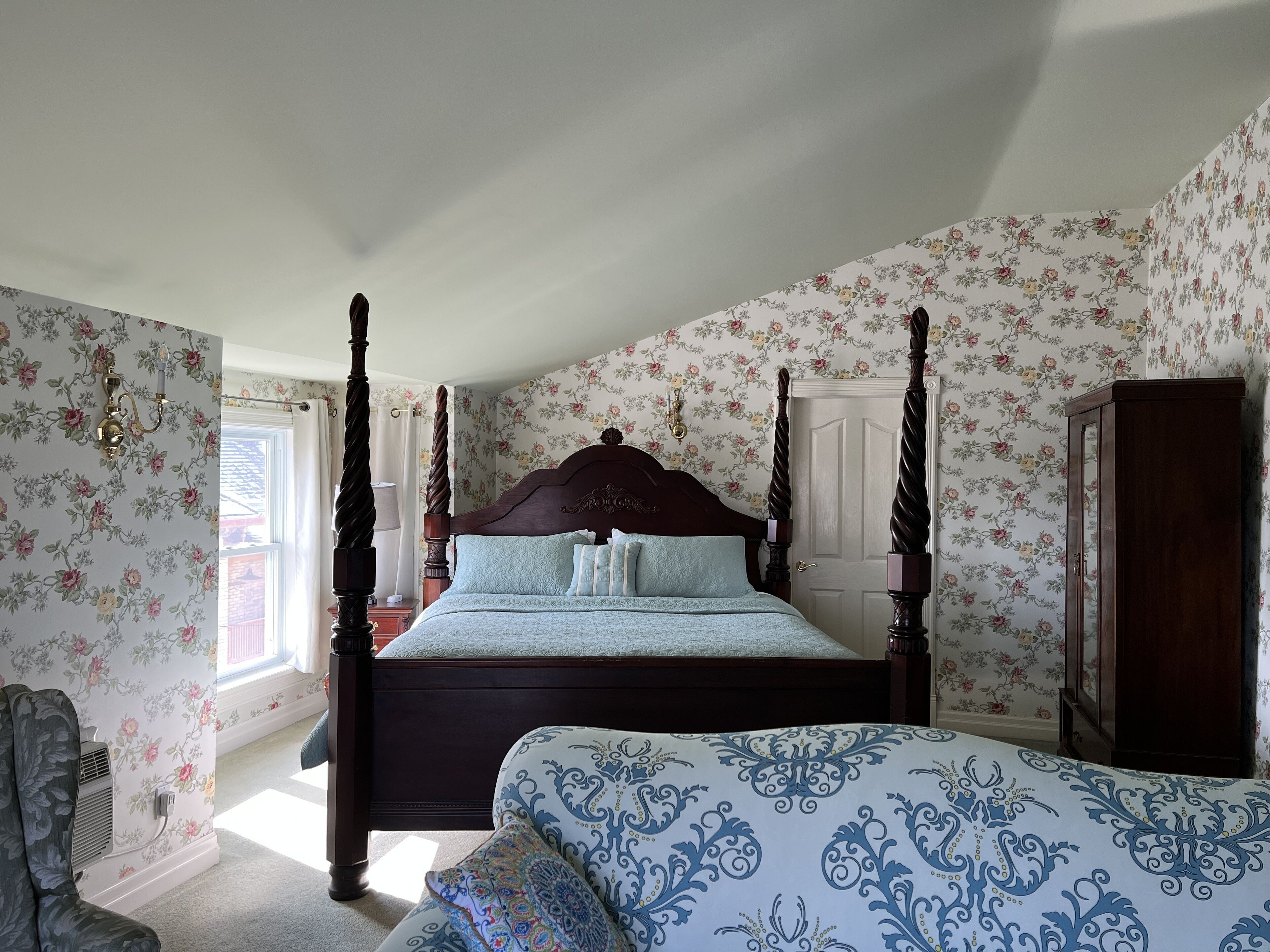 A four-poster bed with floral wallpaper, a wooden wardrobe, and a window with curtains.