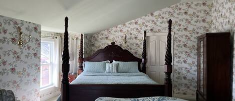 Een hemelbed met bloemenbehang, een houten garderobe en een raam met gordijnen.
