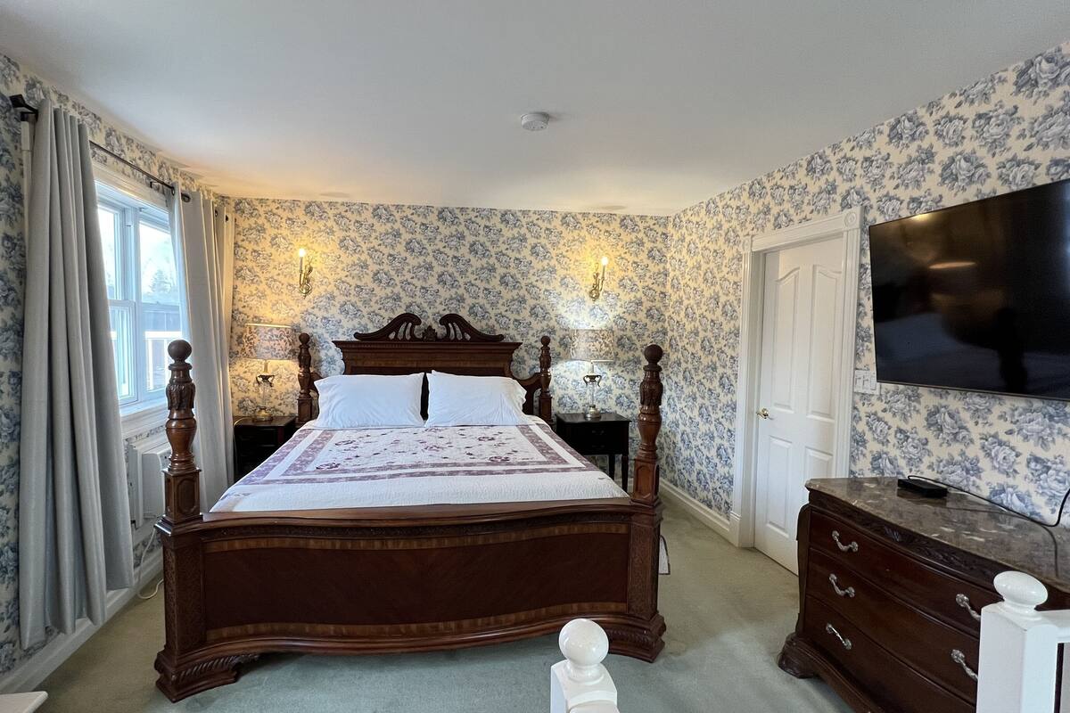 A bedroom with a wooden four-poster bed, floral wallpaper, a television, and a dresser.