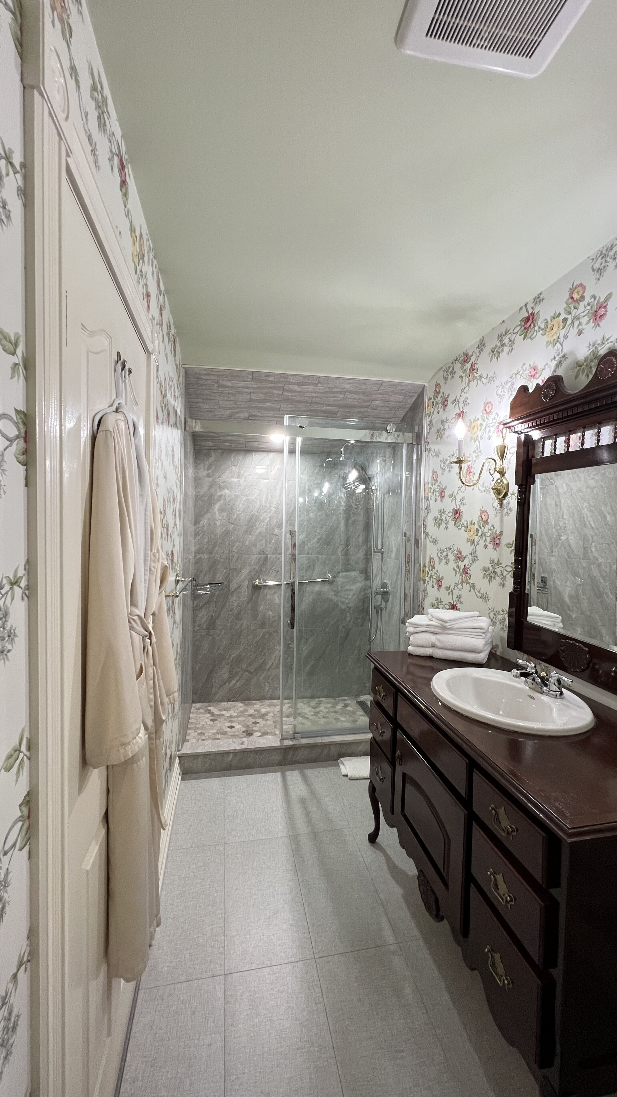A bathroom with a glass-enclosed shower, a vanity with a mirror, and floral wallpaper.