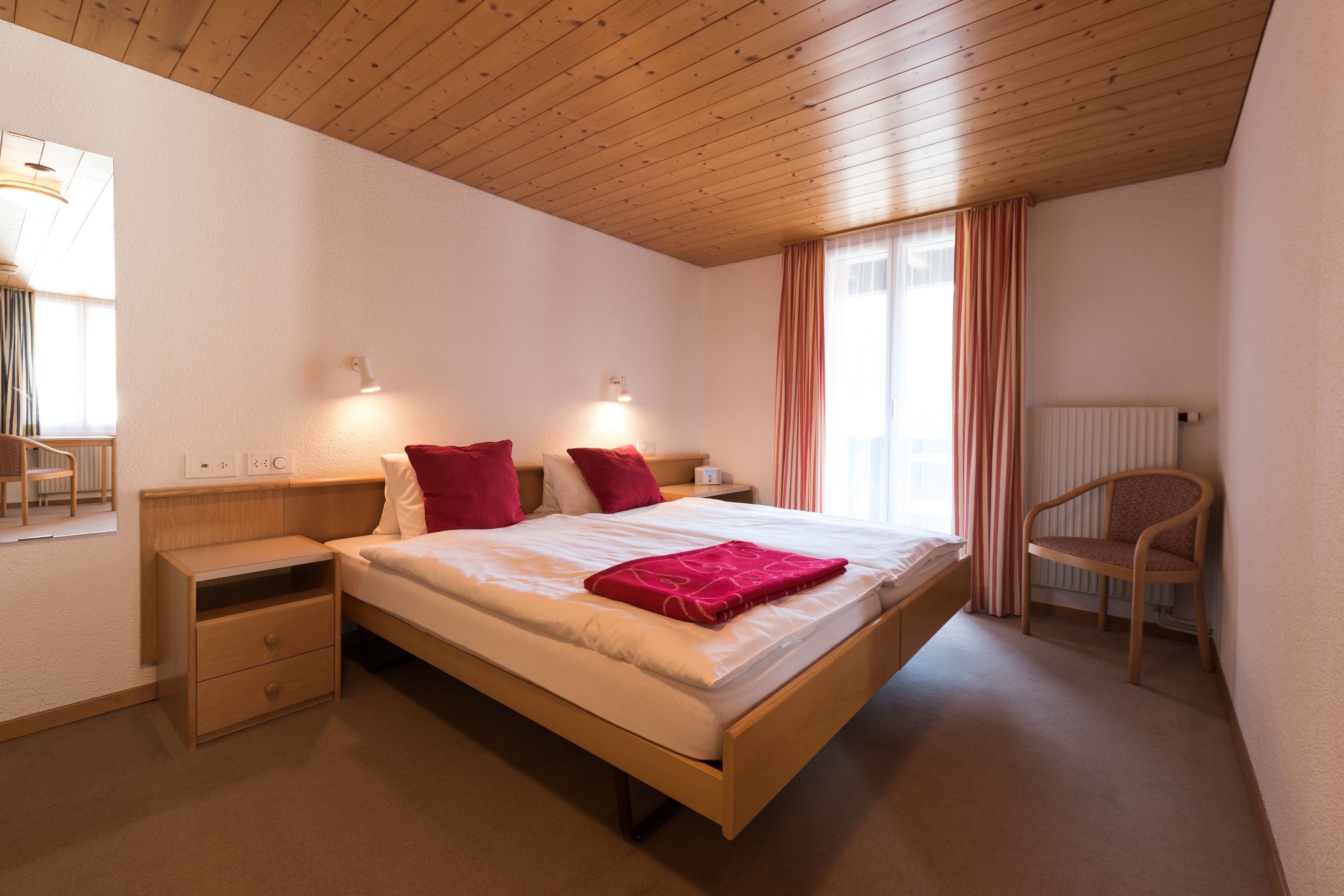A bedroom with a wooden bed, two bedside tables, a chair, and a window with curtains.