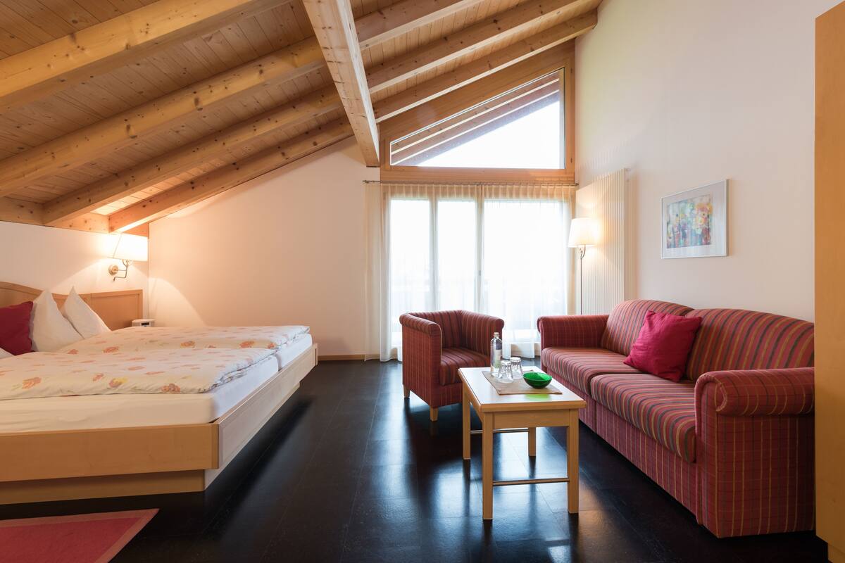 A bedroom with a bed, a sofa, a chair, a table, and a window with curtains.