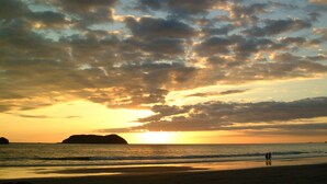 Une scène de plage avec un coucher de soleil, une mer calme et des reliefs terrestres à l’horizon.