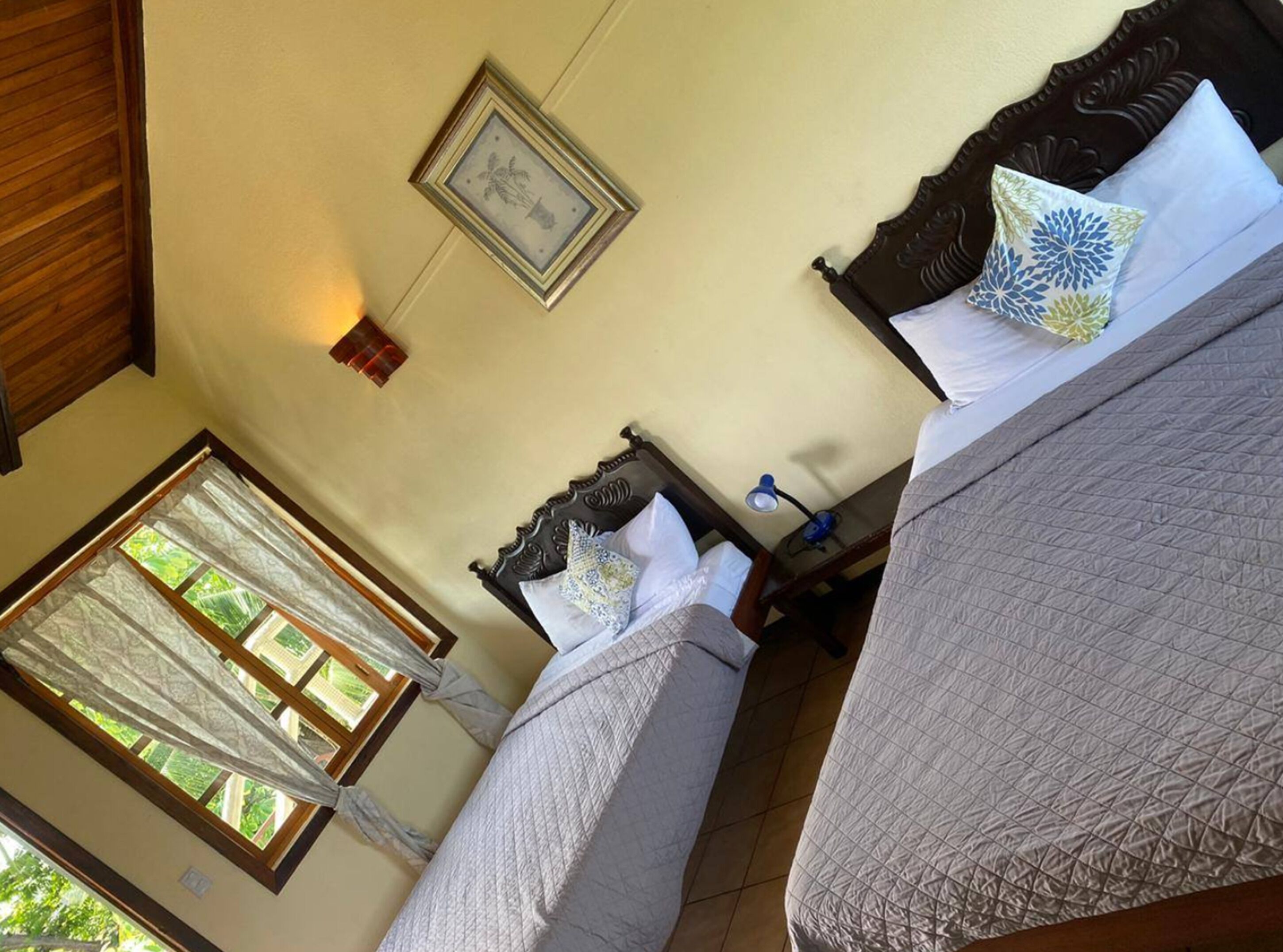 A bedroom with two beds, a window with curtains, a bedside table with a lamp, and a framed picture on the wall.