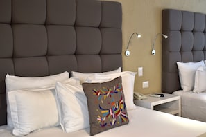 Habitación de hotel con una cama grande, ropa de cama blanca, una almohada colorida, una mesita de noche con un teléfono y un control remoto, y una lámpara de pared.