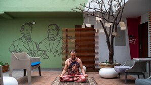 Una persona medita sobre un estrambòs a un espai exterior amb una paret verda, un arbre i mobiliari.