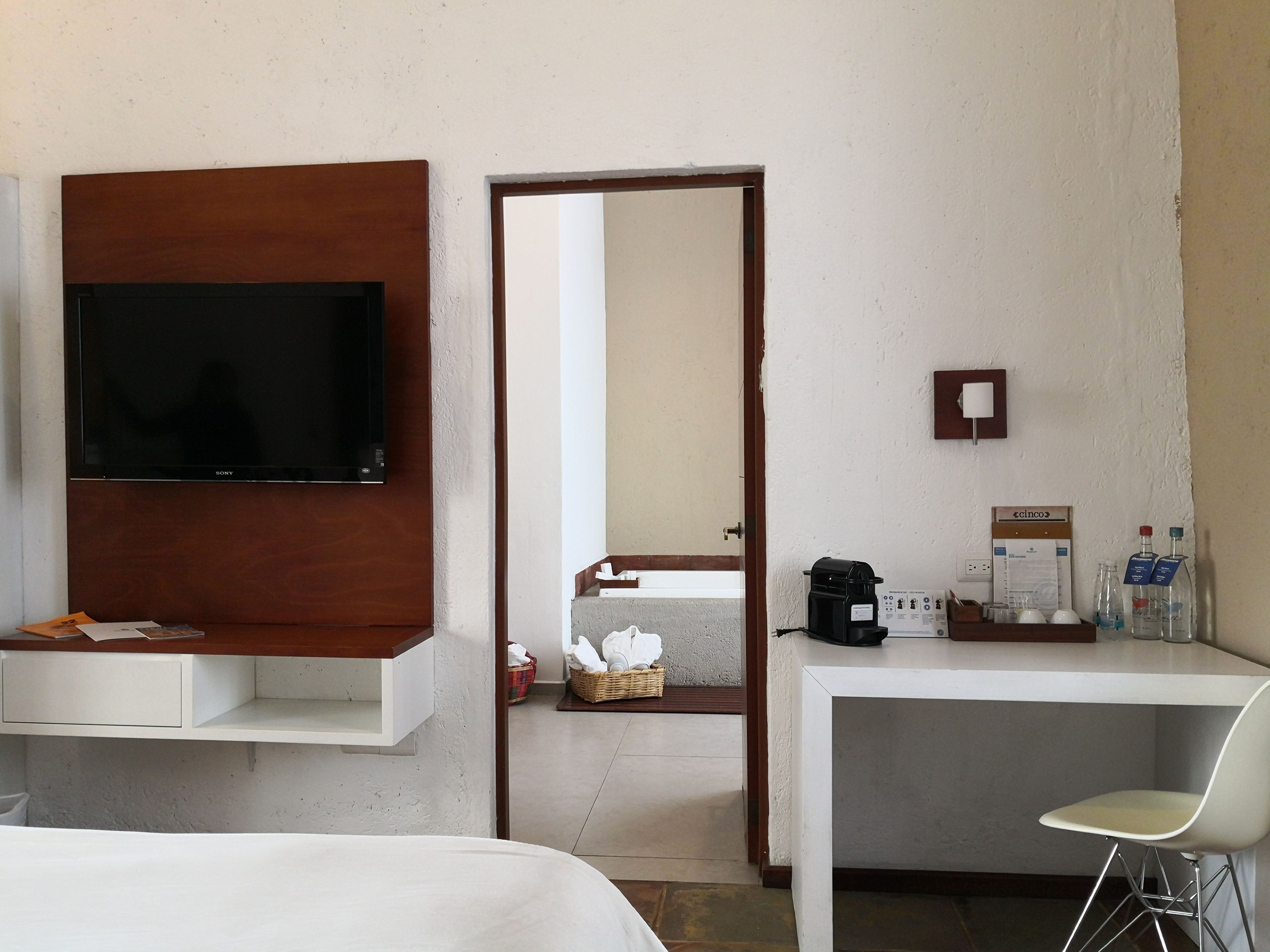 Habitación de hotel con una televisión montada en un panel de madera, un escritorio blanco con una silla, una cafetera y un minibar.