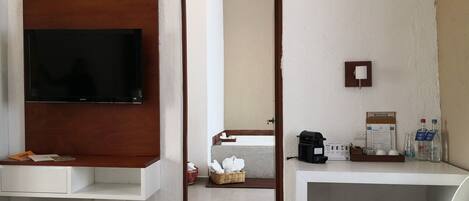 Habitación de hotel con una televisión montada en un panel de madera, un escritorio blanco con una silla, una cafetera y un minibar.