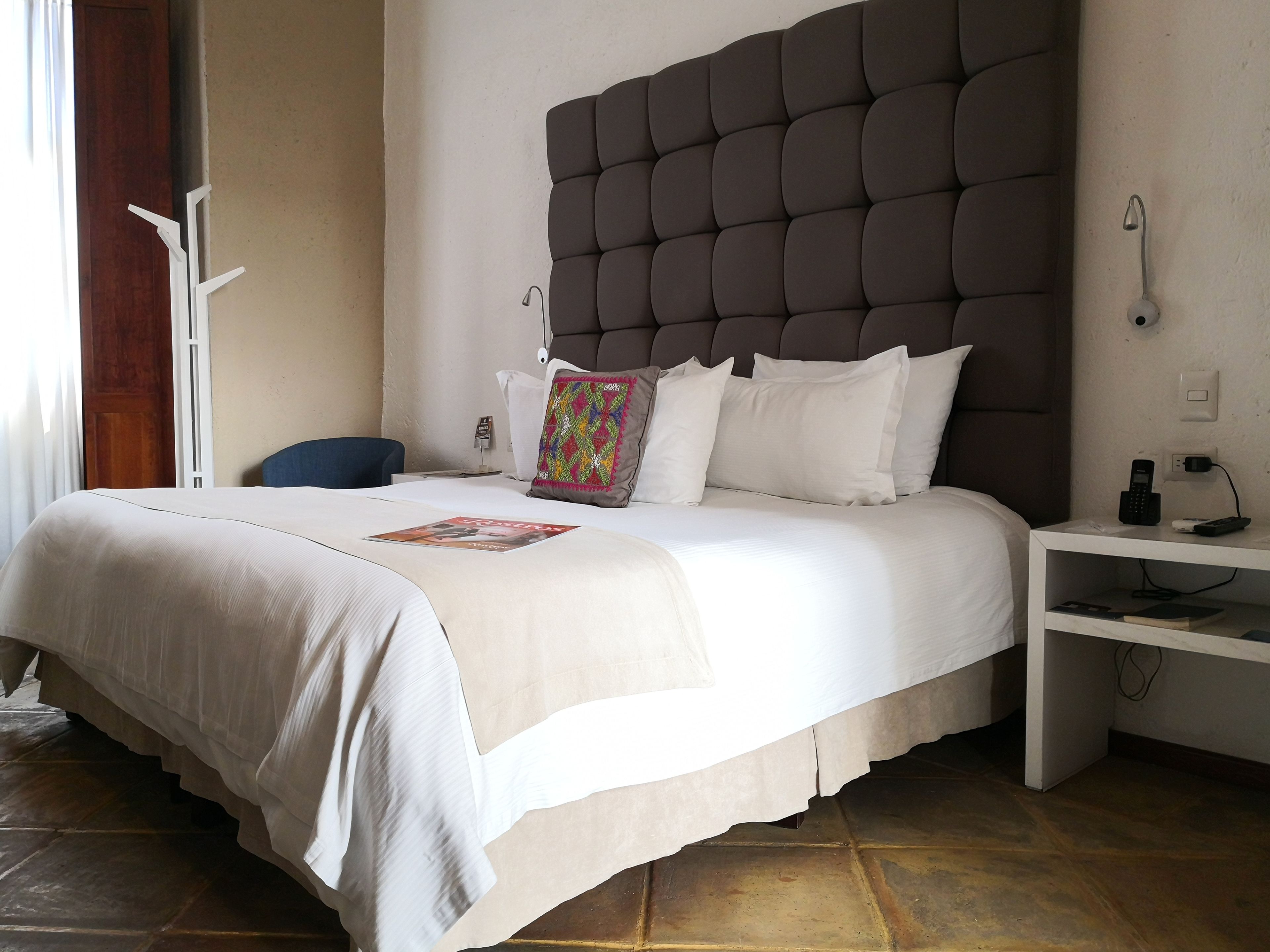 A neatly made bed with a tufted headboard, white bedding, and a colorful pillow. There is a bedside table with a phone and a lamp.